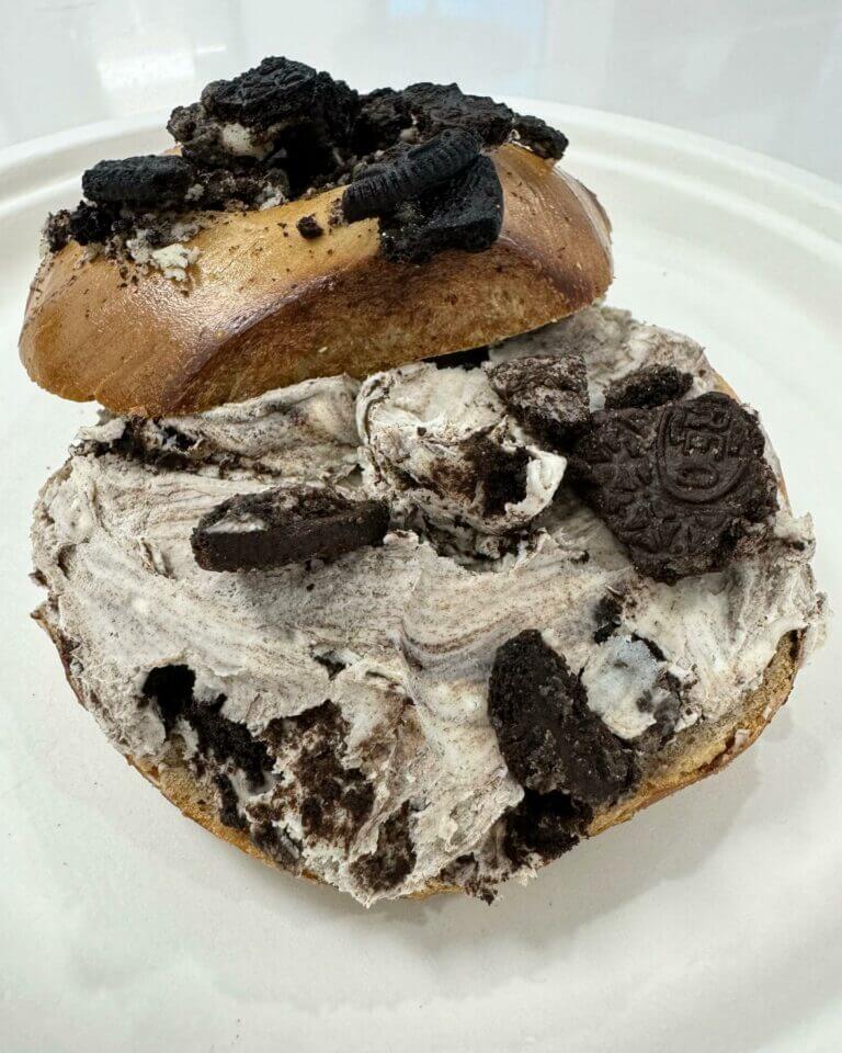 Bagel topped with cookies and cream spread and Oreo pieces on a white plate. Perfect for dessert lovers.