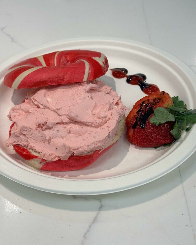 Red and white bagel with strawberry cream cheese, fresh strawberries, and berry sauce on a white plate.