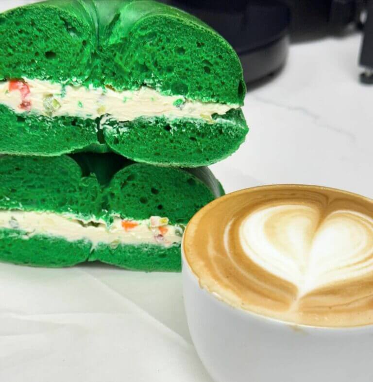 Green bagel with cream cheese and colorful veggies, paired with a latte featuring heart foam art on a white table.