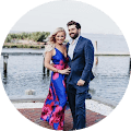 Couple in formal attire by waterfront, woman in floral dress, man in navy suit, scenic outdoor setting.