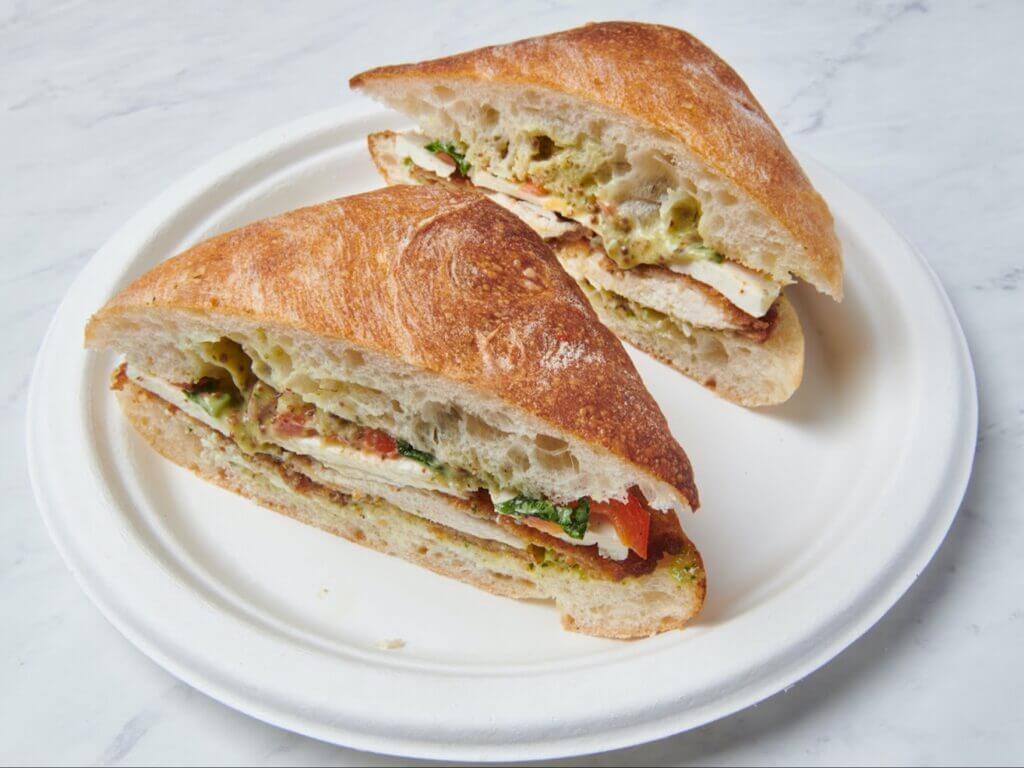 Ciabatta sandwich with grilled chicken, pesto, tomatoes, and mozzarella on a white plate. Fresh, delicious, gourmet lunch option.