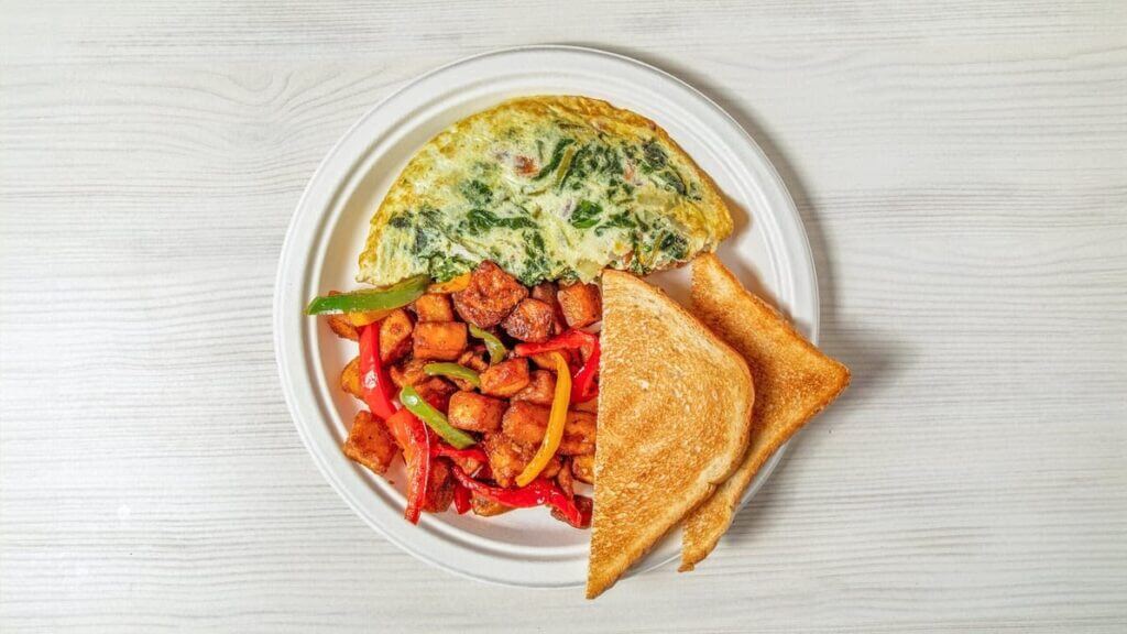 Plate with spinach omelette, roasted potatoes, bell peppers, and toasted bread slices on a white background.