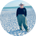 Person in wide-brim hat and green pants stands on sunny beach, ocean in background.